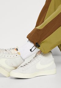 Hvide Nike høj-top sneakers med snørebånd, båret med hvide ribbede sokker og olivengrønne bukser med justerbare snore i benene.