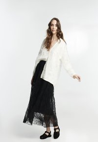 Cardigan blanc texturé avec un col en V et des boutons, porté sur une jupe maxi en dentelle noire ornée de motifs floraux, accompagnée de sandales noires.