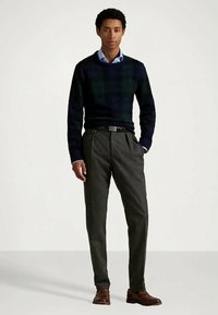 Νεαρός άνδρας στέκεται φορώντας καρό πουλόβερ σε σκούρους τόνους πάνω από ανοιχτό μπλε πουκάμισο με κολάρο, γκρι πλισέ παντελόνι, μαύρη ζώνη και καφέ δερμάτινα παπούτσια.