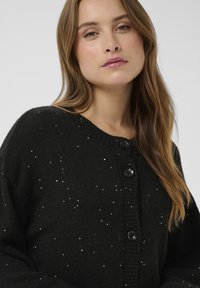 Fekete kötött kardigán bordázott nyakkivágással, négy nagy fekete gombbal és a szöveten szétszórt ezüst díszítésekkel.