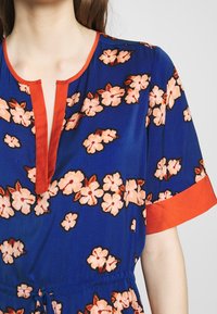 Blauwe bloemenjurk met roze en oranje bloemenpatronen. Heeft een diepe V-hals, oranje bies en korte mouwen. Glad stof,Gathered taille.