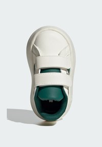 Zapatilla blanca de cuero con dobles correas de Velcro, forro textil verde, forma redondeada y suela ligeramente texturizada.