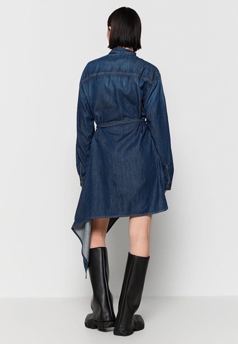 Femme de dos portant une robe en denim bleu foncé asymétrique avec une ceinture et des bottes noires jusqu'aux genoux.