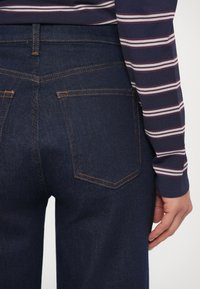 Vaqueros de mezclilla azul oscuro con costuras naranjas, con dos bolsillos traseros, mostrados desde atrás, combinados con un top de manga larga a rayas.