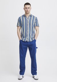 Camicetta a maniche corte con righe verticali blu e beige, abbinata a pantaloni blu. L'outfit include sneakers bianche con dettagli neri.