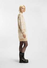 Beige neulemekko, jossa on suuri valkoinen "HUGO" -brändäys, pitkät hihat ja ribatut hihansuut. Yhdistetty mustiin polvimittaisiin saappaisiin.