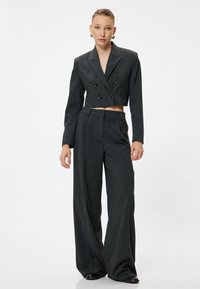 Blazer gris raccourci avec un design croisé et des boutons noirs associé à un pantalon gris taille haute et ample. Texture du tissu lisse.