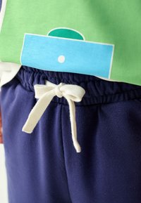 Žalia ir mėlyna grafinė marškinėlių ilgomis rankovėmis sluoksniuota ant tamsiai mėlynų sportinių kelnių su elastine juosmens dalimi ir kreminės spalvos raišteliais. Lygios, kasdienio dėvėjimo audinio tekstūros.