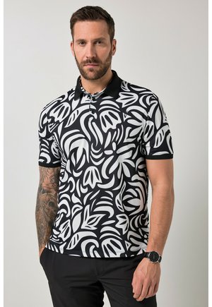 Mann mit Bart, der ein schwarz-weißes Poloshirt mit Blumenmuster, schwarze Hose, Uhr und ein Tattoo-Ärmel am linken Arm trägt, die Hände in den Taschen stehend.