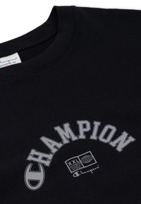 Sudadera negra con cuello acanalado, gran texto en blanco "CHAMPION" en un arco, un logo de globo y "XXL AUTHENTIC APPAREL" debajo.