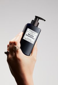 Zwarte rechthoekige fles met baardshampoo met een pompkop, voorzien van een wit etiket met productdetails. 250ml, geproduceerd in Engeland.
