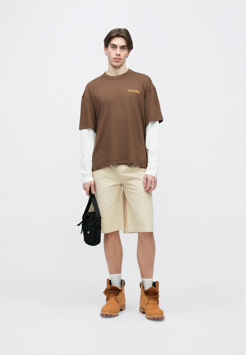 Jeune homme portant un t-shirt marron Dickies par-dessus un sous-pull blanc à manches longues, un short beige jusqu'aux genoux, des bottes de travail couleur sable, et tenant un sac bandoulière noir.