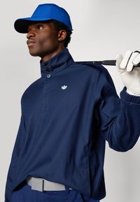 Homem em boné azul e casaco de golfe azul-marinho segura um taco de golfe sobre o ombro, usando uma luva branca, de pé contra um fundo liso.