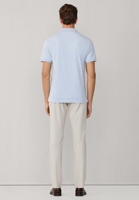 Man die met de rug naar de camera staat, gekleed in een lichtblauw poloshirt met korte mouwen, beige broek en donkerbruine schoenen, voor een effen achtergrond.