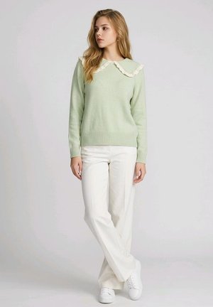 Joven mujer con un suéter verde pálido con un cuello blanco con volantes, pantalones blancos y zapatillas blancas, de pie con las piernas cruzadas.