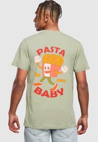 Gaiši zaļa kokvilnas T-krekls ar karikatūras pasta varoni oranžā un sarkanā krāsā, ar uzrakstu "PASTA la vista BABY" biezā fontā.