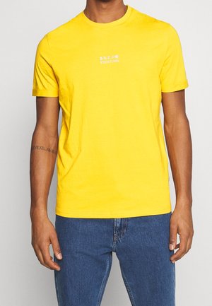 Homme portant un t-shirt jaune vif avec le texte blanc « YOURTURN » et des caractères japonais, associé à un jean bleu, debout devant un fond uni.