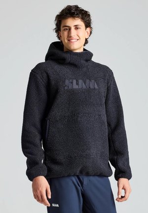 Un hombre sonriente luce una sudadera negra con capucha y textura, y unos pantalones azul marino sobre un fondo liso y claro.
