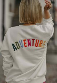 Bílá mikina s barevným vyšitým nápisem "ADVENTURE" v živých barvách. Měkká tkanina s ribbovaným límcem a manžetami, volný střih.