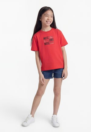 Jente med langt mørkt hår som smiler, iført en rød Moschino T-skjorte, denimshorts og hvite sneakers, står foran en hvit bakgrunn.