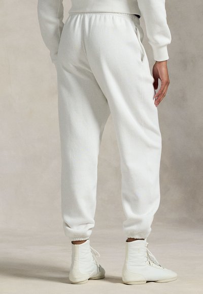 Άτομο που φοράει λευκά παντελόνια φόρμας με ελαστική μέση και λευκά ψηλά αθλητικά παπούτσια, στέκεται με το ένα χέρι δίπλα στο ισχίο του.