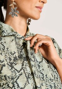 Frau, die den Kragen einer Bluse mit Schlangenprint anpasst, trägt große florale Strassohrringe, gezeigt von Hals bis Hand.