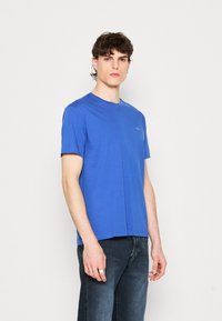 Blaues Baumwoll-T-Shirt mit Rundhalsausschnitt und kurzen Ärmeln, das ein kleines Logo auf der linken Brust zeigt. Getragen mit dunklen Jeans.