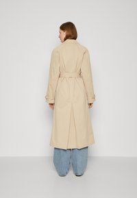 Beige trenchcoat med belte i livet, lange ermer og detaljer med vent bak. Kåpen har en glatt tekstur og en lengde til kneet.