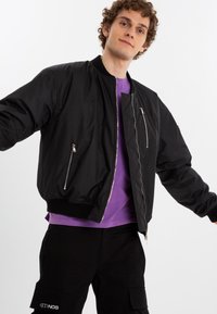 Black bomber jacket with zippered pockets, ribbed cuffs, and a smooth finish. Worn over a purple shirt, modeled against a plain background.