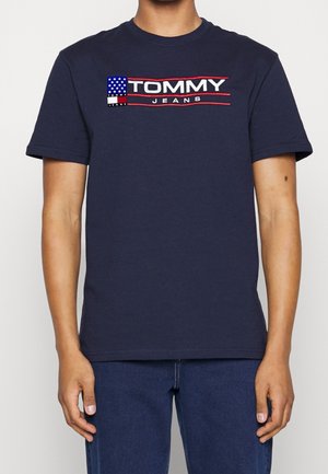 Osoba nosząca granatowy t-shirt Tommy Jeans z haftowaną amerykańską flagą i logo na piersi.