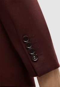 Manica di giacca bordeaux con texture liscia, cinque bottoni marrone scuro con quattro fori ciascuno, dettagli cuciti sui bottoni.