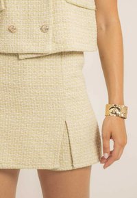 Ensemble deux pièces beige clair en tissu texturé avec des éclats jaunes ; la jupe possède une fente à l'avant, assortie à un bracelet épais en or.