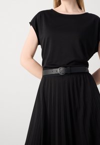 Musta lyhythihainen paita, joka on yhdistetty mustaan laskostettuun hameeseen ja mustaan nahkavyöhön, jossa on pyöreä solki ja säädettävät reiät.