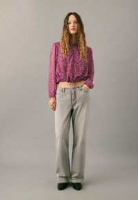 Häkelstil, bauchfreies lila Oberteil mit Kordelzug am Saum; lockere graue Hose mit hoher Taille; schwarze Stiefeletten; Modell steht vor neutralem Hintergrund.