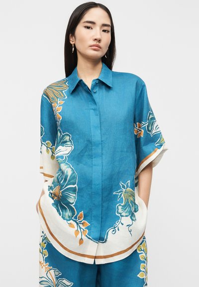 Νεαρή γυναίκα που φοράει μπλε πουκάμισο με μεγάλα λουλουδένια σχέδια και φαρδιά μανίκια, με τα χέρια στις τσέπες, στέκεται μπροστά σε ένα απλό φόντο.