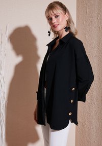 Cămașă neagră cu nasturi, cu o croială lejeră, având nasturi aurii pe lateral și guler. Purtată peste un top alb.
