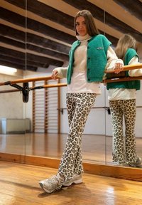 Gebreide groene vest over een witte coltrui, gecombineerd met een flared broek in luipaardprint en lichtgrijze sneakers, staat in een spiegelende studio.