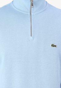 Ljusblå zip-up tröja gjord av ribbat material. Har en hög krage och en liten broderad krokodil-logotyp på bröstet. Dragkedjedetaljen är synlig.