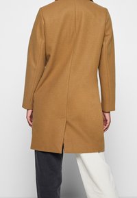 Brun kappa i ullblandning med rak silhuett, långa ärmar och slits vid fållen. Slät textur och minimalistisk design.