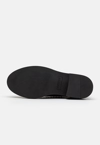 Semelle de chaussure noire avec grip texturé, talon rectangulaire, et nom de marque "TWINSET" embossé près de la zone de la voûte plantaire sur un fond neutre.