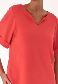 Haut corail à manches courtes avec un col en V et des manches retroussées, fabriqué en tissu doux ; présente une texture lisse et une coupe décontractée.