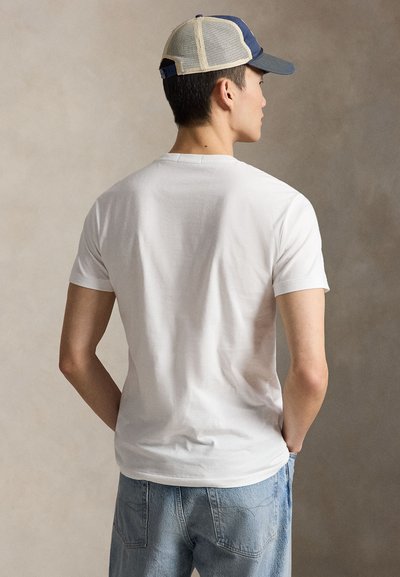 Ung mann vendt bort fra oss iført en hvit kortermet T-skjorte, lyseblå jeans og en mørkeblå og beige netting baseballcaps.