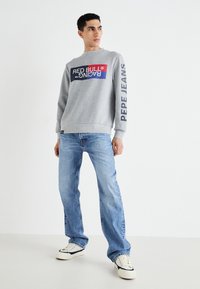 Sivi dukserica s šarenim logotipom "Red Bull Racing" na prsima, dugih rukava i oznakom "Pepe Jeans" na rukavu. Uparena sa svijetloplavim trapericama.