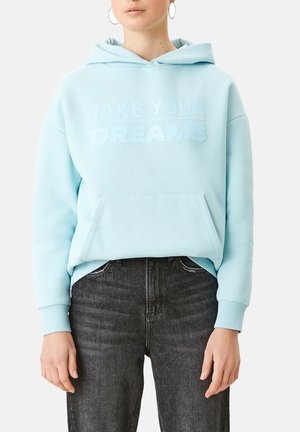 Personne portant un sweat à capuche bleu clair avec le texte "PRENDS TES RÊVES AU SÉRIEUX" et un jean noir, debout devant un fond blanc.