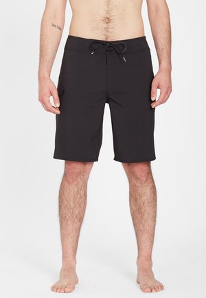 Shorts de bain noirs jusqu'aux genoux avec poches latérales et taille à cordon, portés par un homme torse nu debout sur un fond uni.