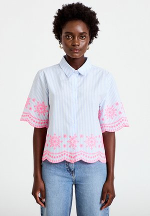 Femme portant une chemise à manches courtes à rayures bleu clair avec broderies florales roses sur les manches et l'ourlet, associée à un jean bleu.