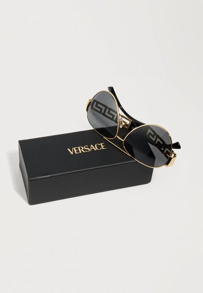 Zwart-gouden pilotenbril met Grieks sleutelmotief op de pootjes, rustend op een zwarte Versace-etui met gouden letters.