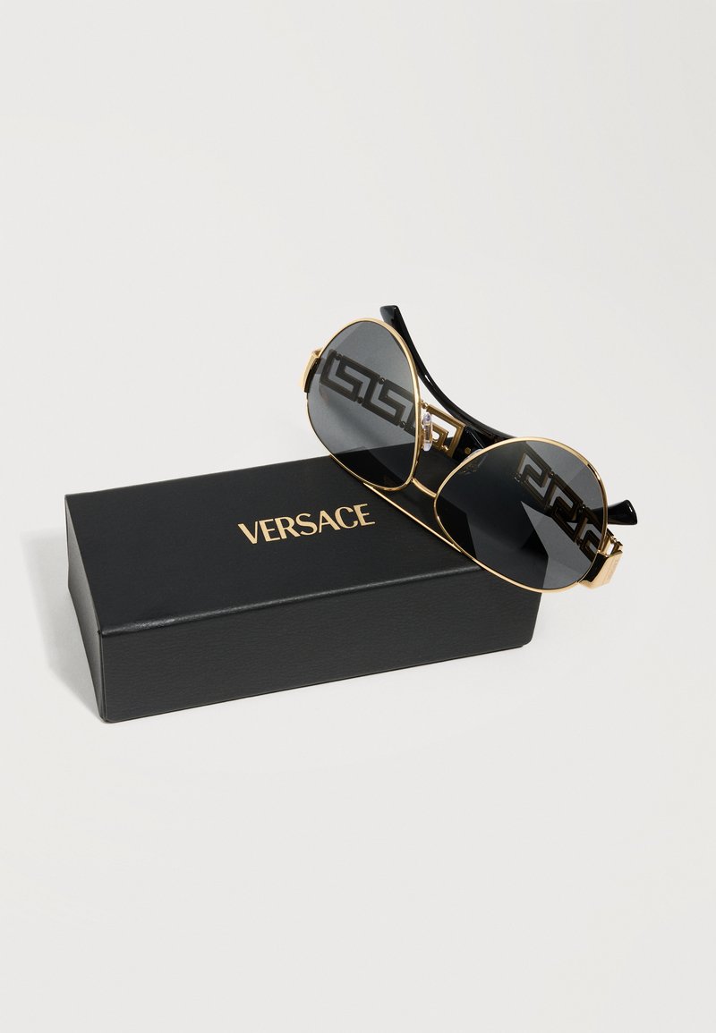 Lunettes de soleil aviateur noires et dorées avec un motif à clé grecque sur les branches, reposant sur un étui Versace noir avec des lettres dorées.