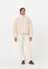 Chaqueta acolchada beige con cuello alto, fabricada en un tejido suave y texturizado. Cierre frontal con cremallera, bolsillos laterales y logotipo en el pecho.