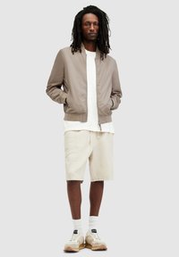 Lätt taupe bomberjacka, vit lager-på-lager t-shirt, beige shorts, vita strumpor och avslappnade beige sneakers med bruna accenter.